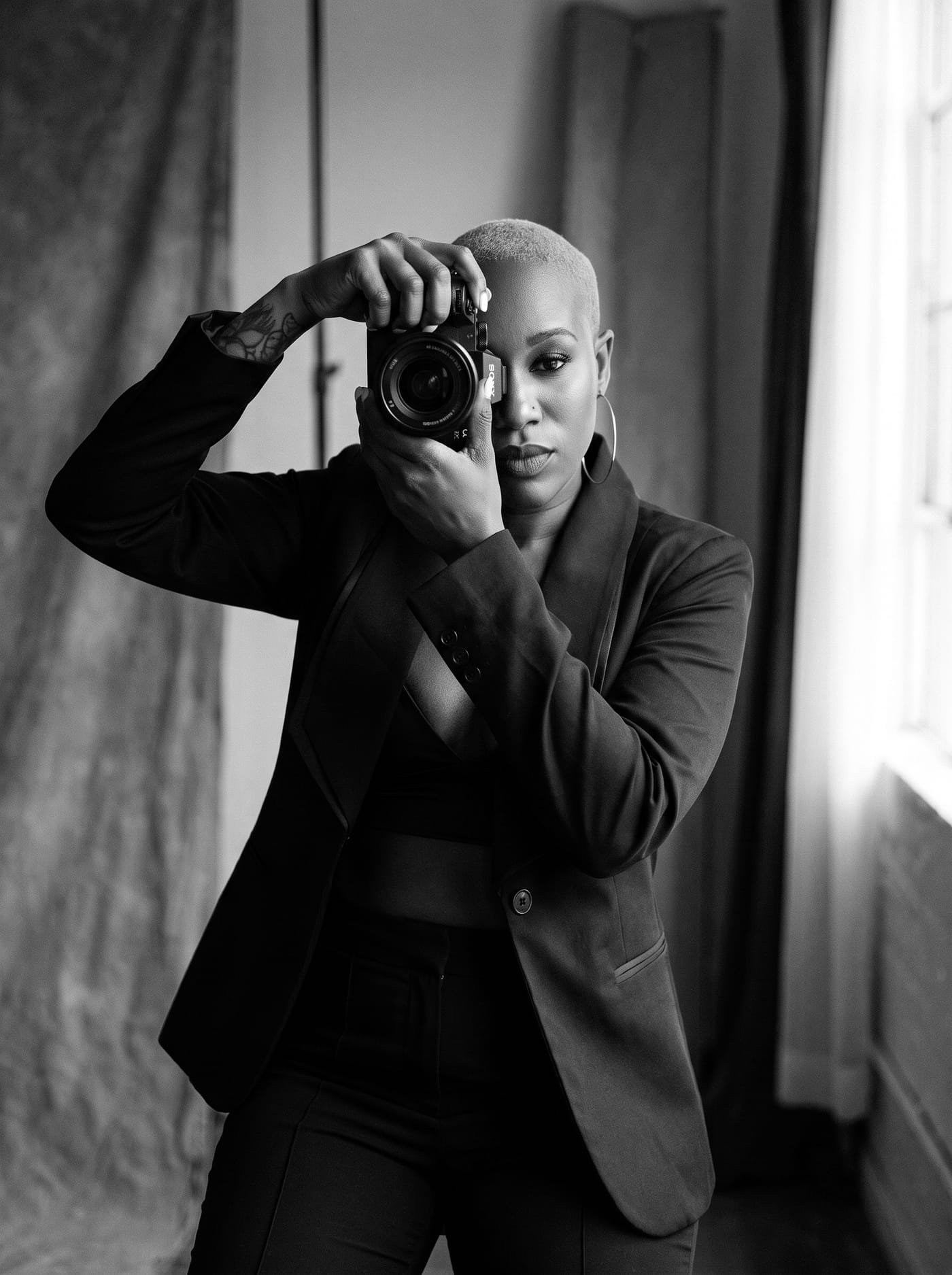 Black and white portrait of Nikki Rumph standing at a window with her camera raised