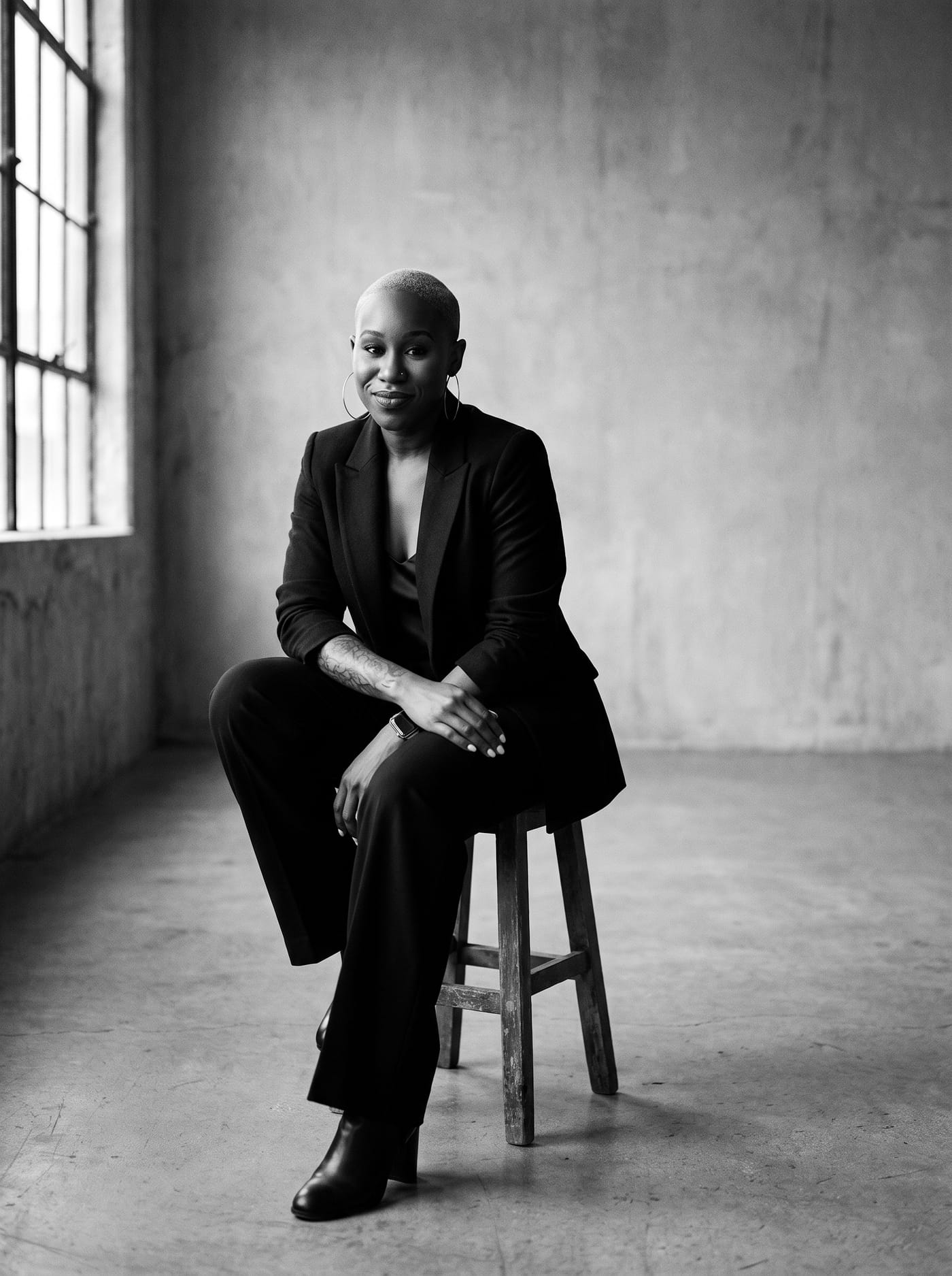 Black and white portrait of Nikki Rumph seated on a wooden stool in an industrial loft beside a large window