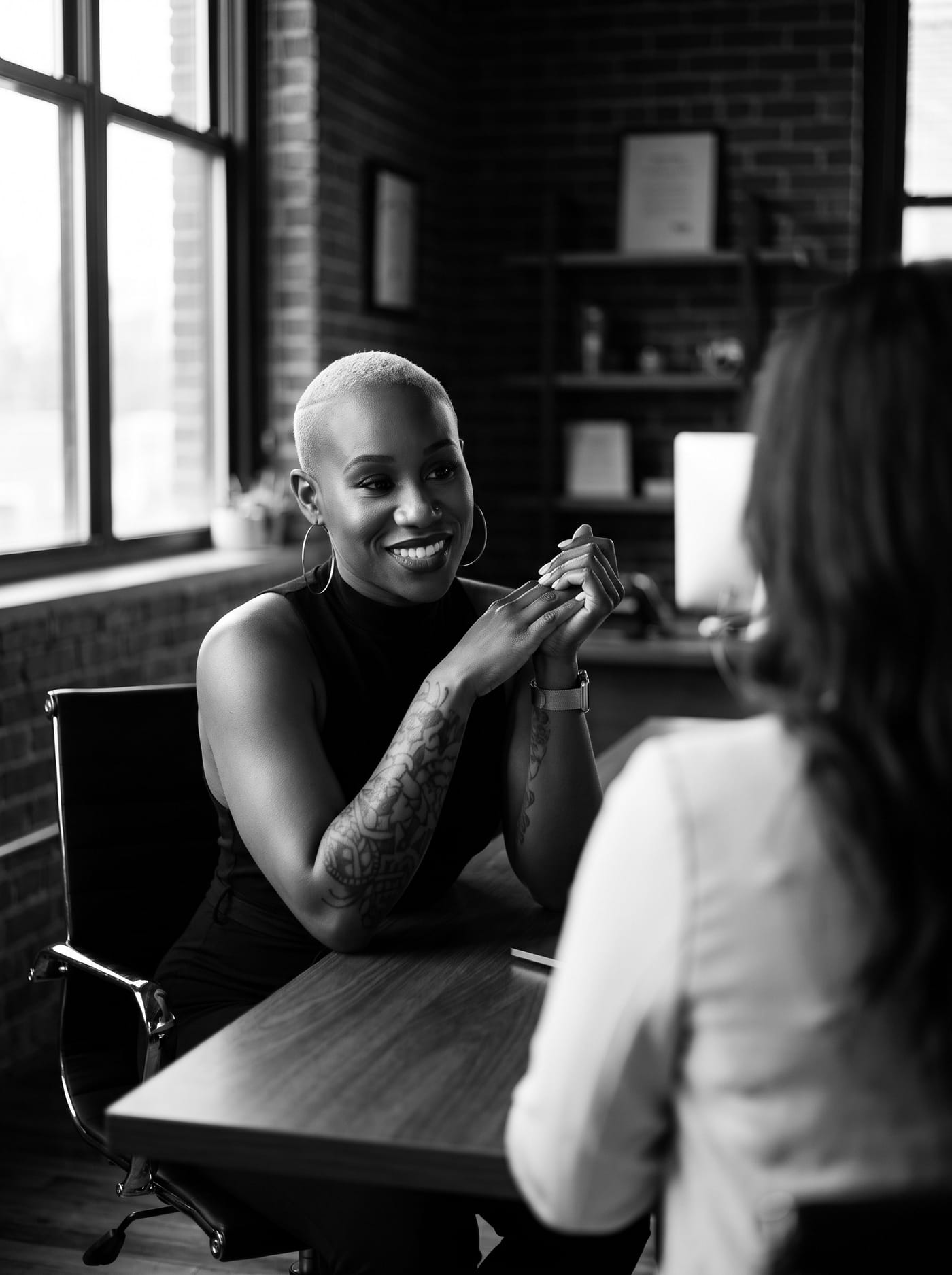 Black and white portrait of Nikki Rumph smiling in conversation with a client across a table in a modern loft office