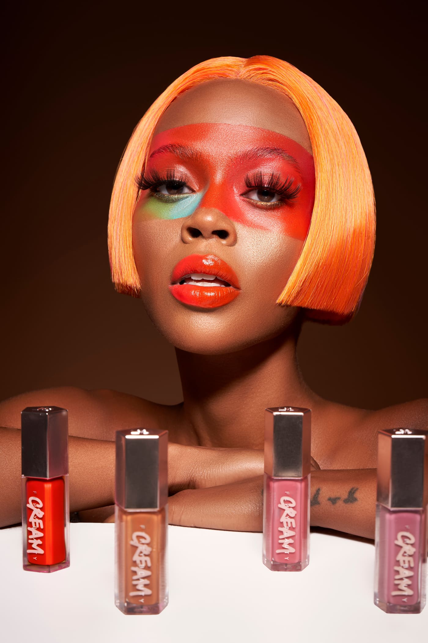 Color beauty editorial of a model with an orange bob and graphic makeup above a row of CREAM lip glosses