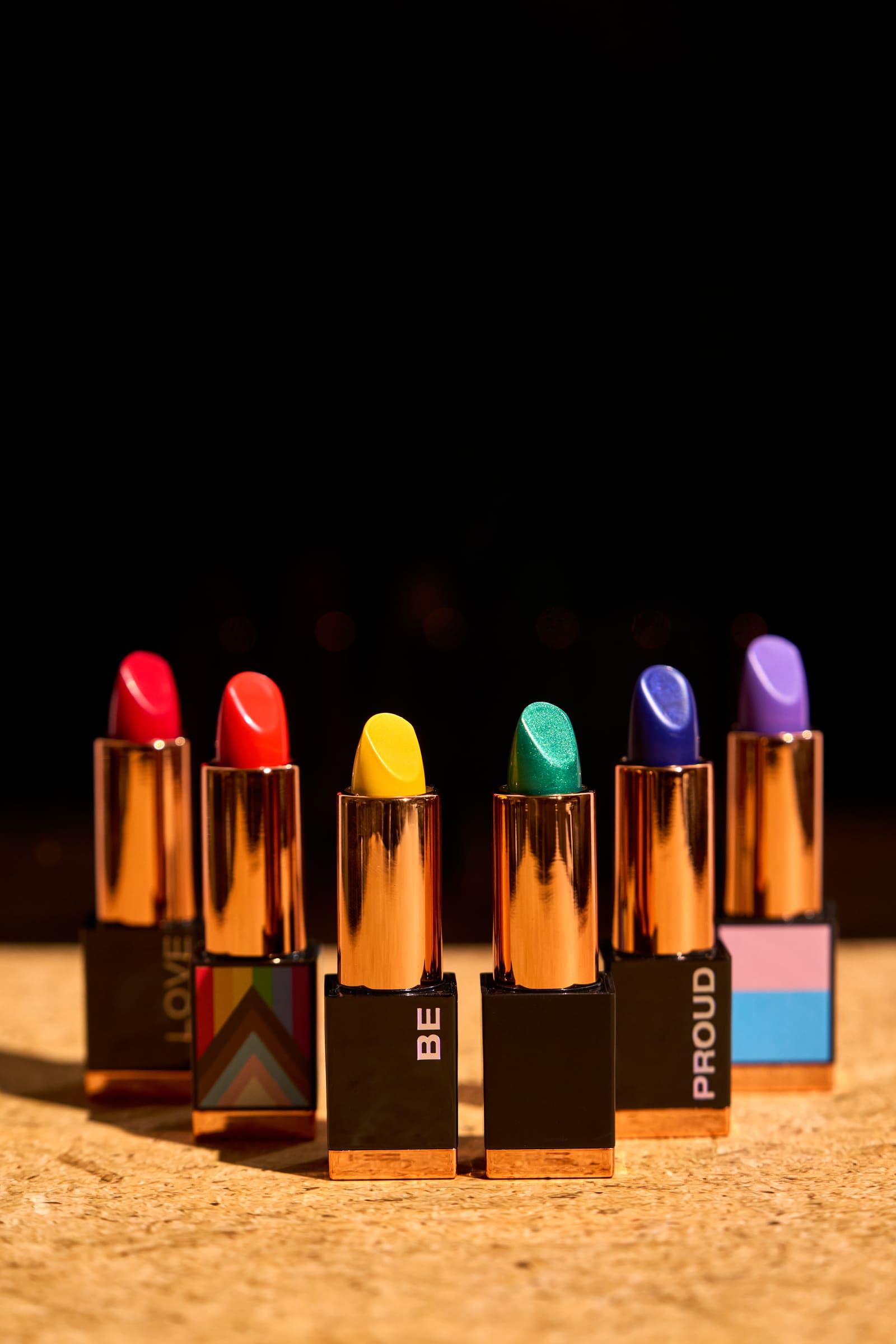 Color product shot of six Kandi Koated Cosmetics pride lipsticks in red, orange, yellow, green, blue and purple on a cork surface